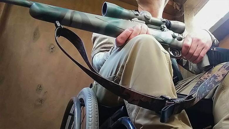 Hunter holding a camouflaged scoped rifle while in a wheelchair in a hunting blind.