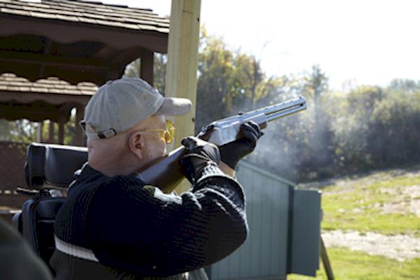 Man in a tan baseball cap shooting a shotgun from a wheelchair.